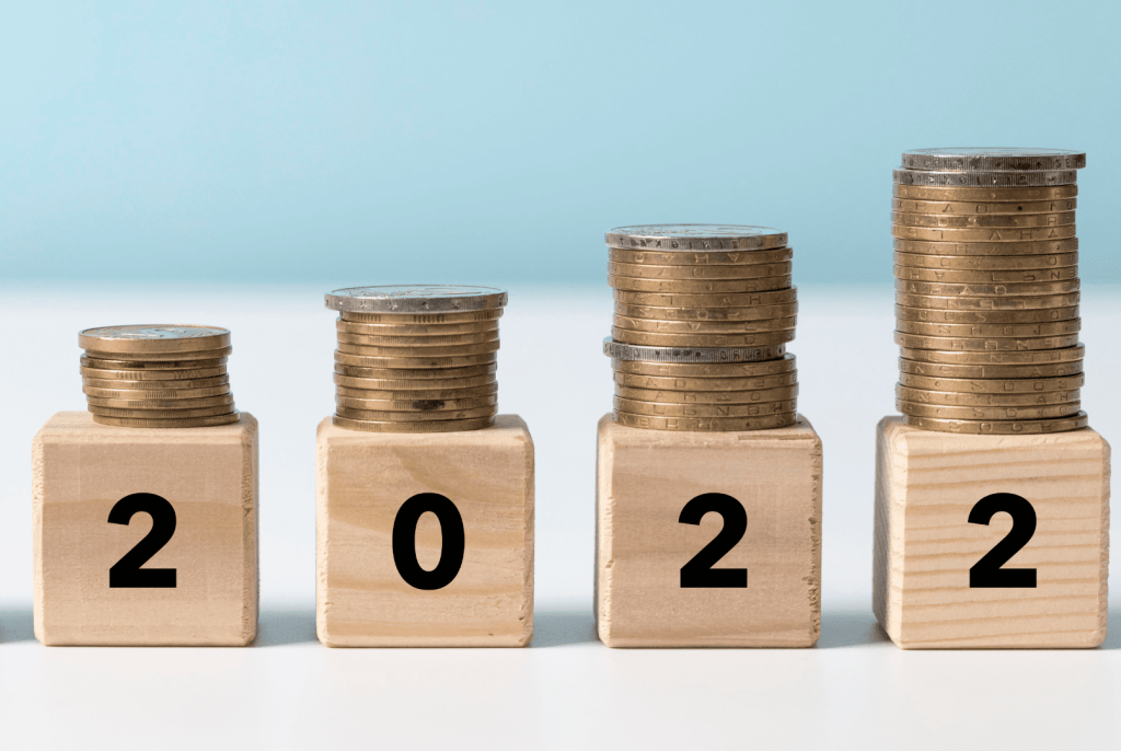 Zdjęcie jest ilustracją artykułu: Rosnąca presja płacowa w firmach – co na ten sądzą temat ekonomiści? Przedstawia drewniane kostki z cyframi 2022 a na nich stosy monet.