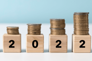 Zdjęcie jest ilustracją artykułu: Rosnąca presja płacowa w firmach – co na ten sądzą temat ekonomiści? Przedstawia drewniane kostki z cyframi 2022 a na nich stosy monet.