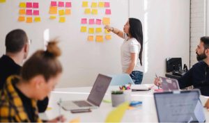 Zdjęcie jest ilustracją artykułu "Jak Agile może pomóc HR?". Przedstawia spotkanie grupy roboczej pracowników. Jedna kobieta stoi przy tablicy pełnej kolorowych post it. Pozostałe osoby z zespołu siedza przy stole konferencyjnym|