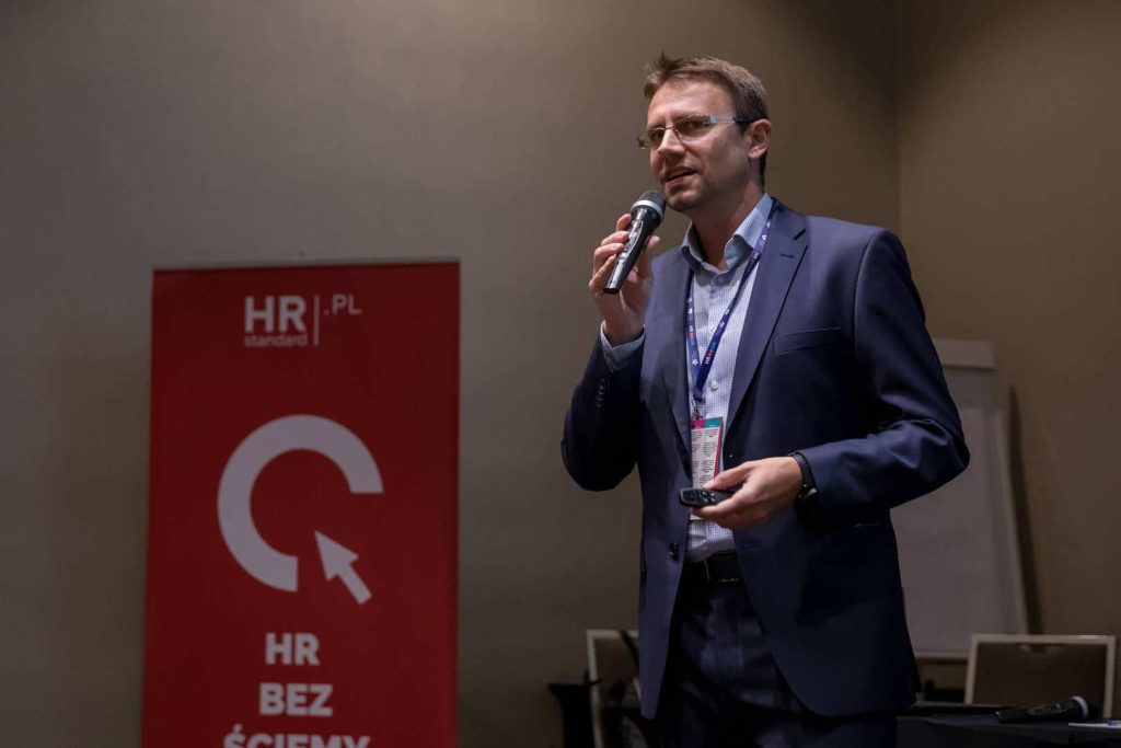 Zdjęcie ilustruje artykuł: X edycja HRcamp za nami! Przedstawia mężczyznę z mikrofonem podczas publicznego wystąpienia.|||