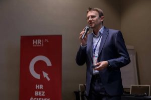 Zdjęcie ilustruje artykuł: X edycja HRcamp za nami! Przedstawia mężczyznę z mikrofonem podczas publicznego wystąpienia.|||